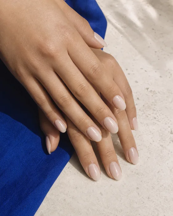 Duas mãos com unhas amendoadas em gel nude leitoso sobre tecido azul e pedra clara